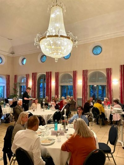 Hoher Saal mit rundem Kristalllüster und roten Vorhängen; an runden Tischen sitzen etwa dreißig Personen, in der Wand über den Fenstern runde blaue Zierfenster.