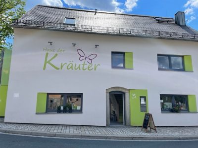 Weiß verputztes Haus mit grün gerahmten Fenstern und dem großen Schriftzug „Haus der Kräuter“ samt Schmetterling an der Giebelseite; vor der Tür steht eine Aufstelltafel.