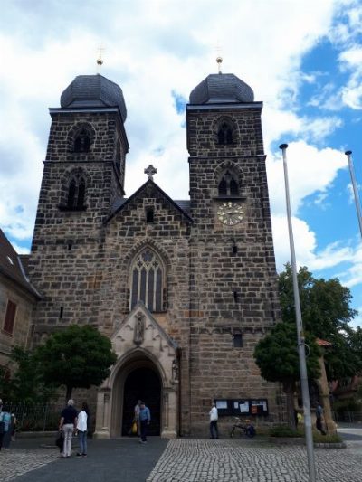 Kirche aus dunklem Sandstein mit zwei Türmen unter geschweiften Hauben; über dem spitzbogigen Portal ein Maßwerkfenster, am rechten Turm eine Uhr. Vor dem Eingang stehen einzelne Personen.
