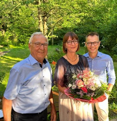 Drei Personen stehen im sonnigen Garten vor dichtem Grün und blicken in die Kamera; die Frau in der Mitte hält einen großen Blumenstrauß.