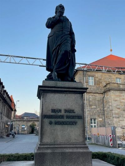 Bronzestandbild eines Mannes im langen Mantel auf hohem Steinsockel mit der Inschrift „Jean Paul Friedrich Richter“; dahinter Sandsteinbauten mit rotem Dach unter blauem Himmel.