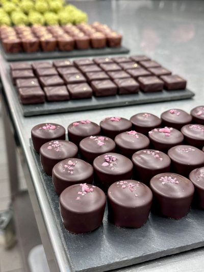 Mehrere Schieferplatten mit Reihen frisch überzogener Pralinen auf einem Edelstahltisch; die vorderen sind mit rosa Streuseln bestreut.
