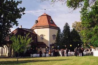Barockes Schlösschen mit geschweifter Haube und Dachreiter, von alten Bäumen umstanden; davor stehen auf der Wiese festlich gedeckte Tische, an denen sich Gäste versammelt haben, dazwischen bunte Lampions.