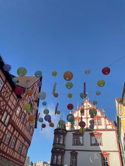 Blick nach oben in eine Altstadtgasse: über der Straße hängen an Drähten viele bunte Lampions, links ein Fachwerkhaus, rechts ein weiß-rotes Bürgerhaus mit geschwungenem Giebel vor blauem Himmel.