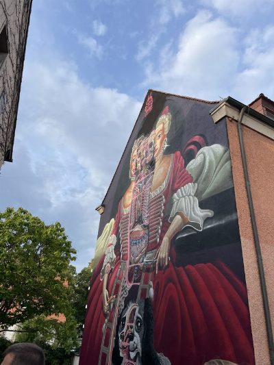 Großes Wandbild an einer Hausgiebelwand: eine Frau in rotem Barockkleid, deren Körper zur Hälfte anatomisch geöffnet gemalt ist und Schädel, Rippen und Organe zeigt; unten ein ebenso dargestellter Hund.