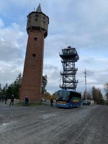 Alter Wasserturm aus Backstein mit umlaufender Aussichtsgalerie, daneben ein moderner Stahlgitterturm mit verglaster Kanzel; davor parkt ein blauer Reisebus, einzelne Personen stehen auf dem Platz.