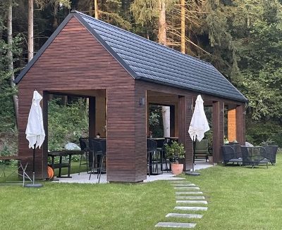 Offener Holzpavillon mit dunklem Satteldach am Waldrand; darunter Stehtische und Barhocker, davor zwei geschlossene weiße Sonnenschirme und Korbsessel. Über den Rasen führt ein Weg aus Trittplatten.