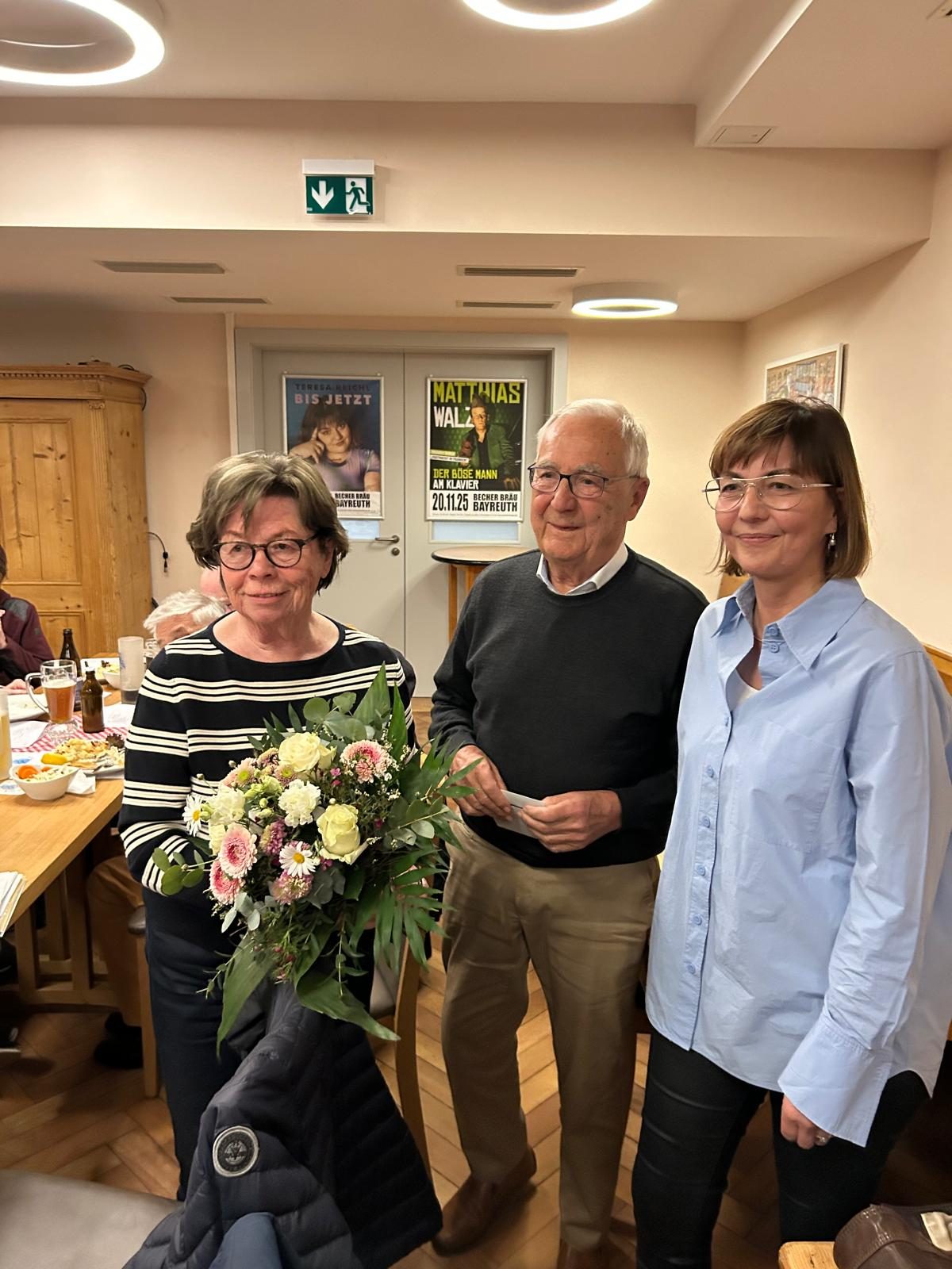 Drei Personen stehen nebeneinander in einem Gasthausraum und blicken in die Kamera; die Frau links hält einen großen Blumenstrauß, der Mann in der Mitte eine Karte. Im Hintergrund ein gedeckter Tisch und Veranstaltungsplakate.