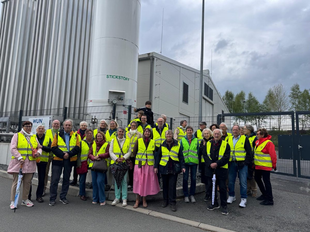 Rund dreißig Personen in gelben Warnwesten stehen dicht beieinander vor einem Industriegelände; dahinter ein hoher Tank mit der Aufschrift „Stickstoff“ und eine Werkshalle.