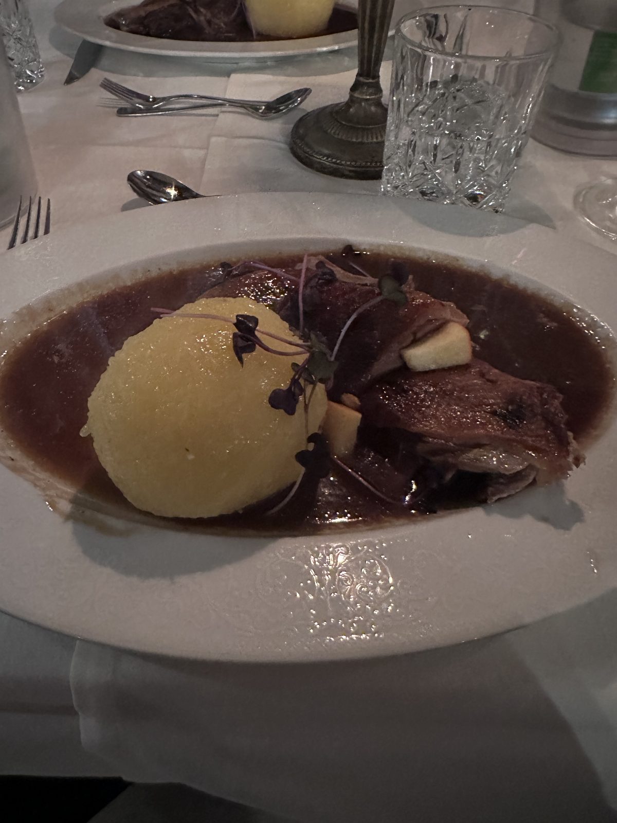 Angerichteter Teller auf weißer Tischdecke: Fleisch in dunkler Soße mit einem Kloß und feinen Kräutern, daneben geschliffene Gläser und Besteck.