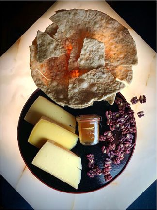 Dunkler Teller mit drei Käsestücken, einem Glas Chutney und einer Handvoll gezuckerter Nüsse; darüber liegt ein großes, dünn gebackenes Fladenbrot.