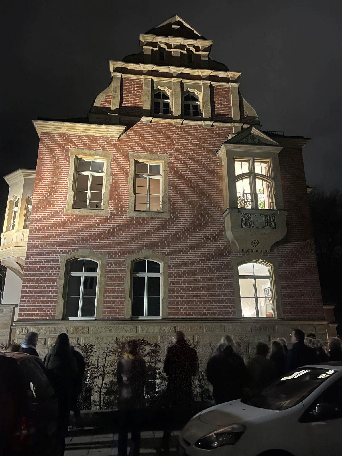 Backsteinvilla bei Nacht, von unten angestrahlt; helle Sandsteingewände, ein geschwungener Giebel und ein Erker, mehrere Fenster sind erleuchtet. Davor stehen einige Personen am Straßenrand.