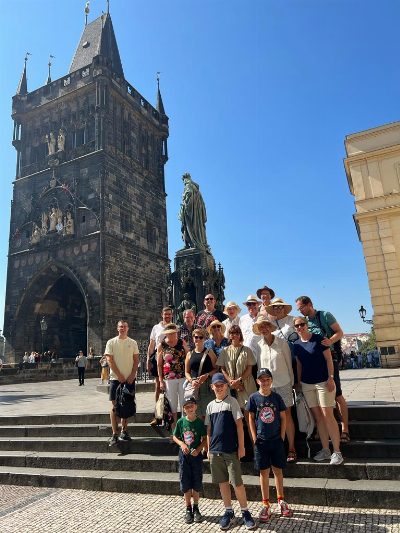 Reisegruppe auf einer Treppe vor einem gotischen Brückenturm aus dunklem Stein; daneben ein steinernes Standbild vor wolkenlosem Himmel.