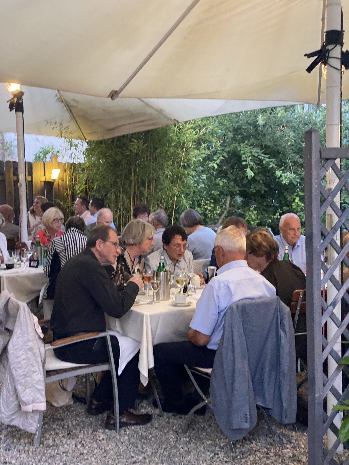 Abendliche Gartenrunde unter einem Sonnensegel: an einer langen weiß gedeckten Tafel sitzen etwa fünfzehn Personen, dahinter dichtes Grün und Lichterketten.