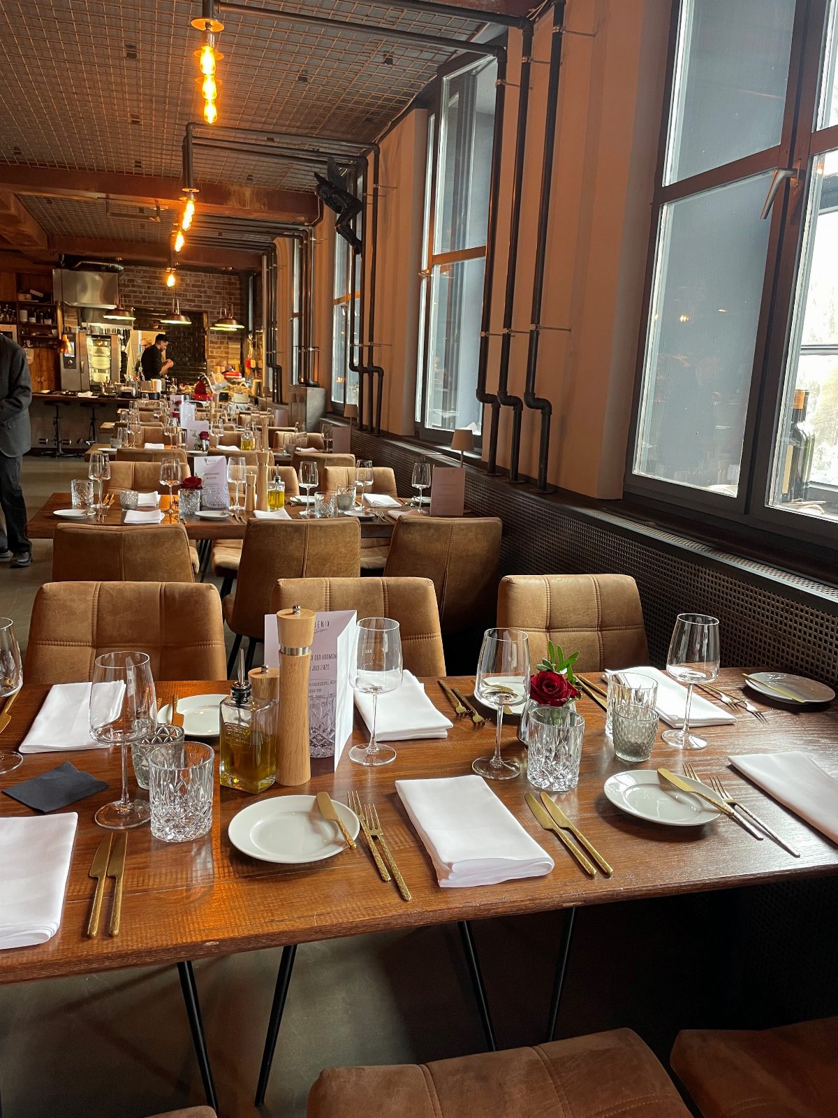 Lange festlich eingedeckte Tafel in einem Restaurant mit Ziegelwänden und offen verlegten Rohrleitungen; braune Lederstühle, weiße Servietten und eine rote Rose als Tischschmuck.