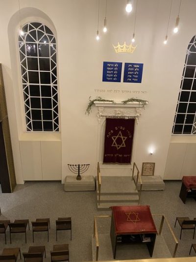 Heller Synagogenraum von der Empore aus: der Toraschrein mit rotem Vorhang und Davidstern, darüber die Gesetzestafeln in hebräischer Schrift und eine goldene Krone; links ein siebenarmiger Leuchter, davor Stuhlreihen und ein Lesepult.